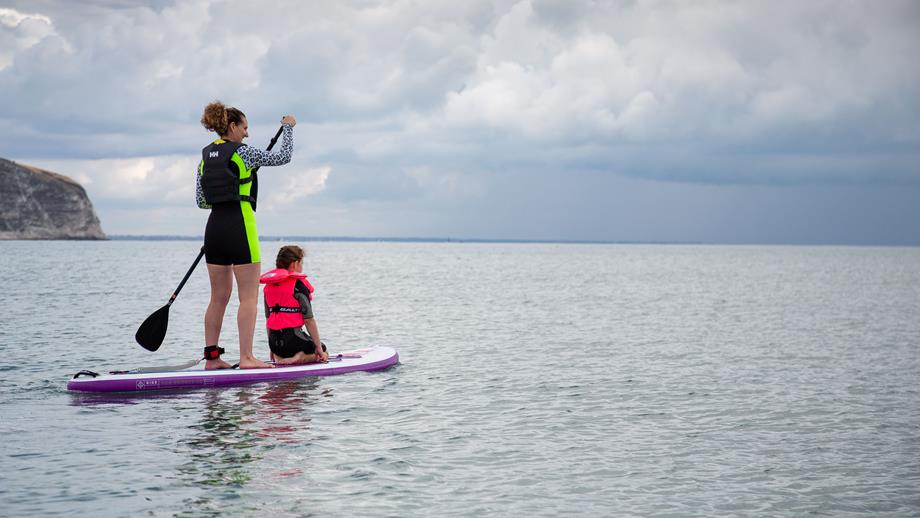A lady and a child wearing buoyancy aids are paddleboarding in a calm, flat sea