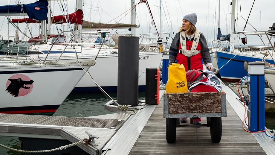 A female sailor who is wearing a lifejacket is pushing a cart full of sailing equipment along a pontoon, with boats moored on either side.