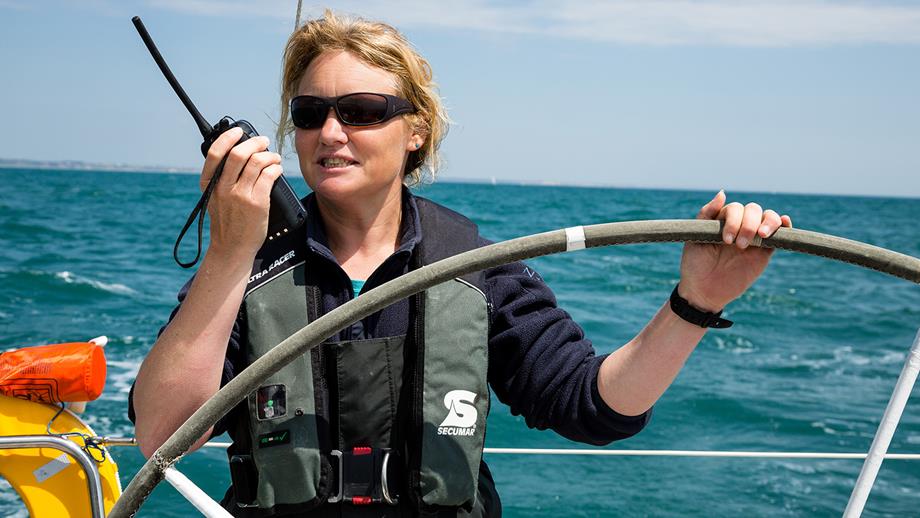 Pip Hare is on a yacht out at sea on a sunny day, wearing a lifejacket and speaking into a VHF radio.