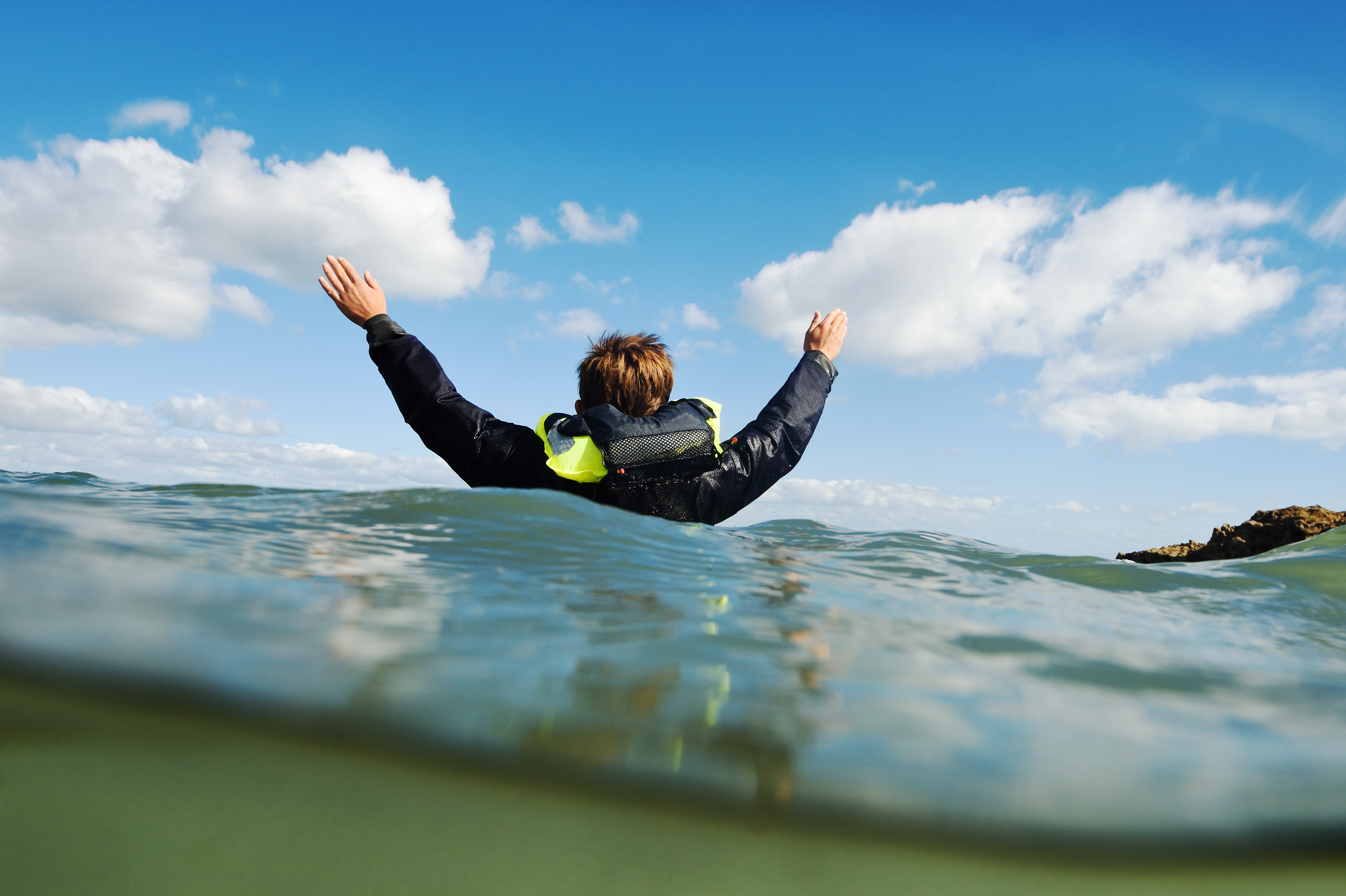A man is in the sea, holding both arms up and wearing a buoyancy aid.