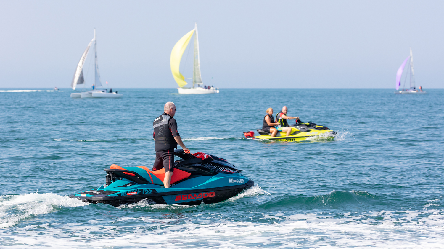 A man is on a personal watercraft on the sea, with another personal watercraft and three sailing vessels in the background