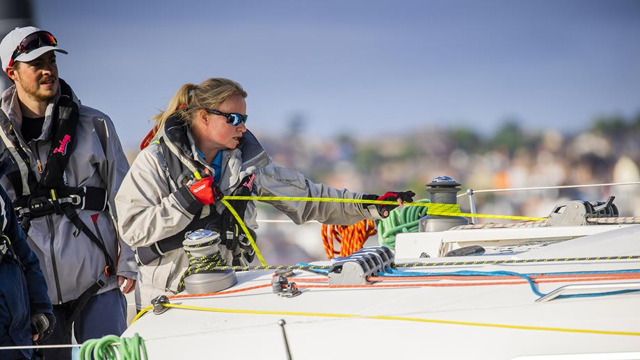 A blonde woman onboard a boat, wearing a grey jacket, sunglasses and a lifejacket, pulls on a florescent yellow rope