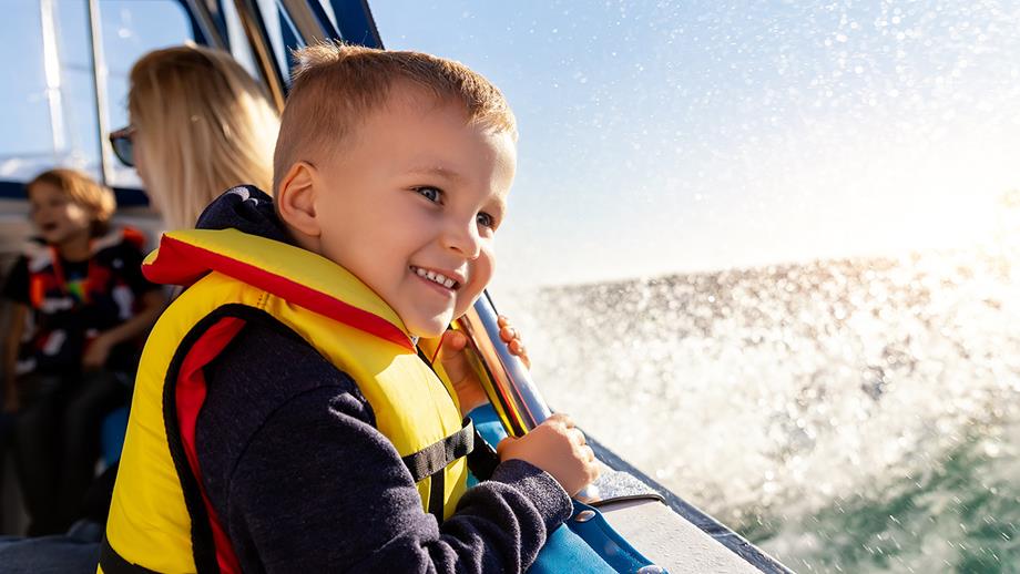 A small blonde boy wearing a yellow lifejacket grins while looking out at the waves from inside a sailing boat