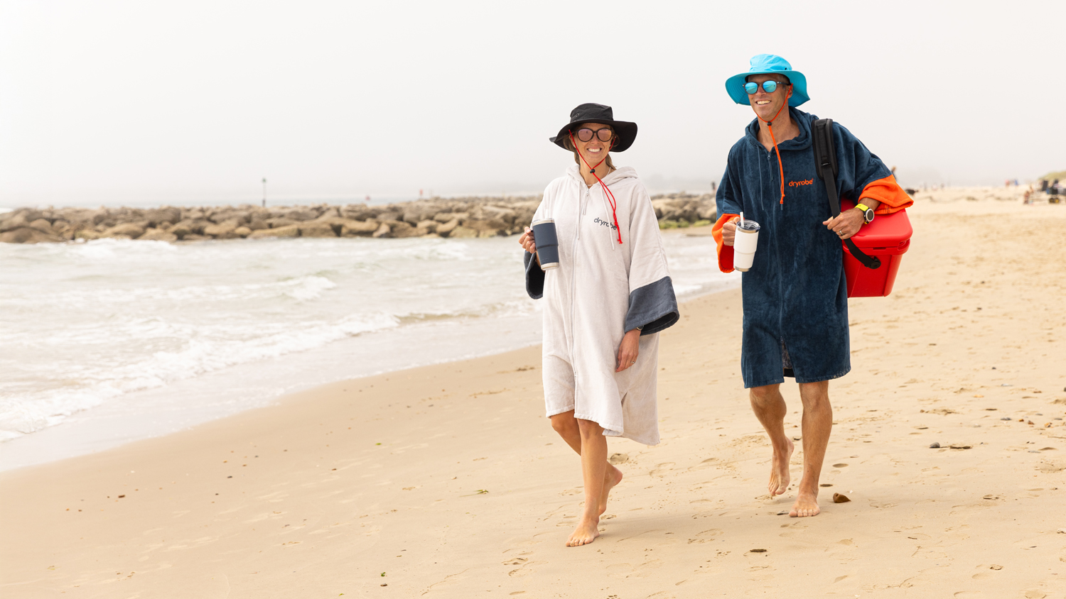 Two smiling people casually walk barefoot along a beach along a beach in dryrobe towel robes and hats and each holding a yeti mug. One is carrying a yeti cool box over their shoulder.