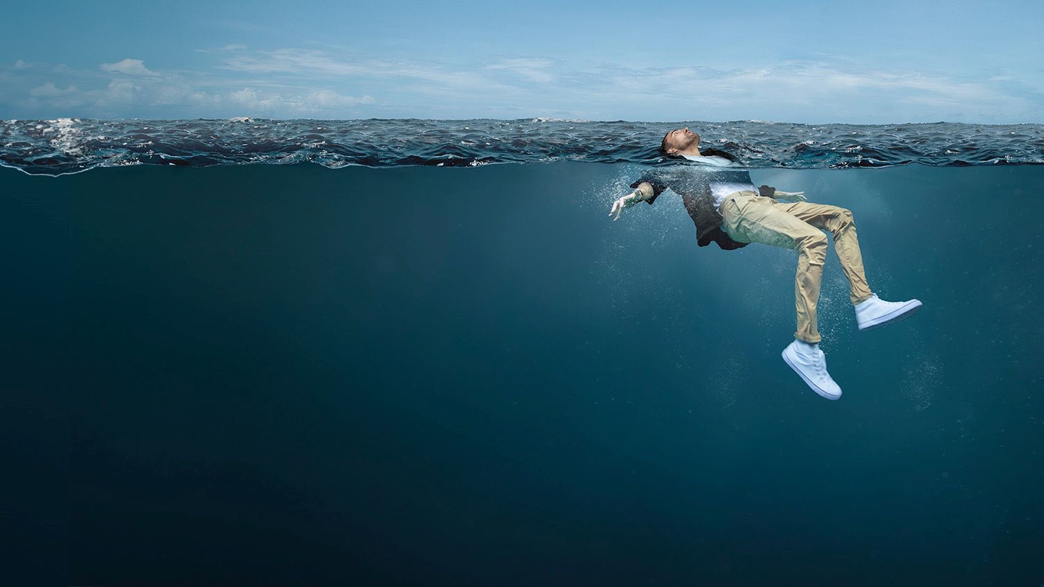 A young man is fully clothed in the open water. His face is above the water, looking up to the sky, and his arms and legs are stretched out, gently moving to help him stay afloat. 