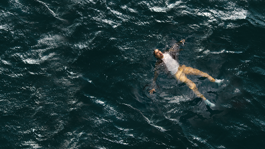 A man wearing a loose shirt, tshirt, trousers and shoes floating on his back, in the sea, with arms and legs
