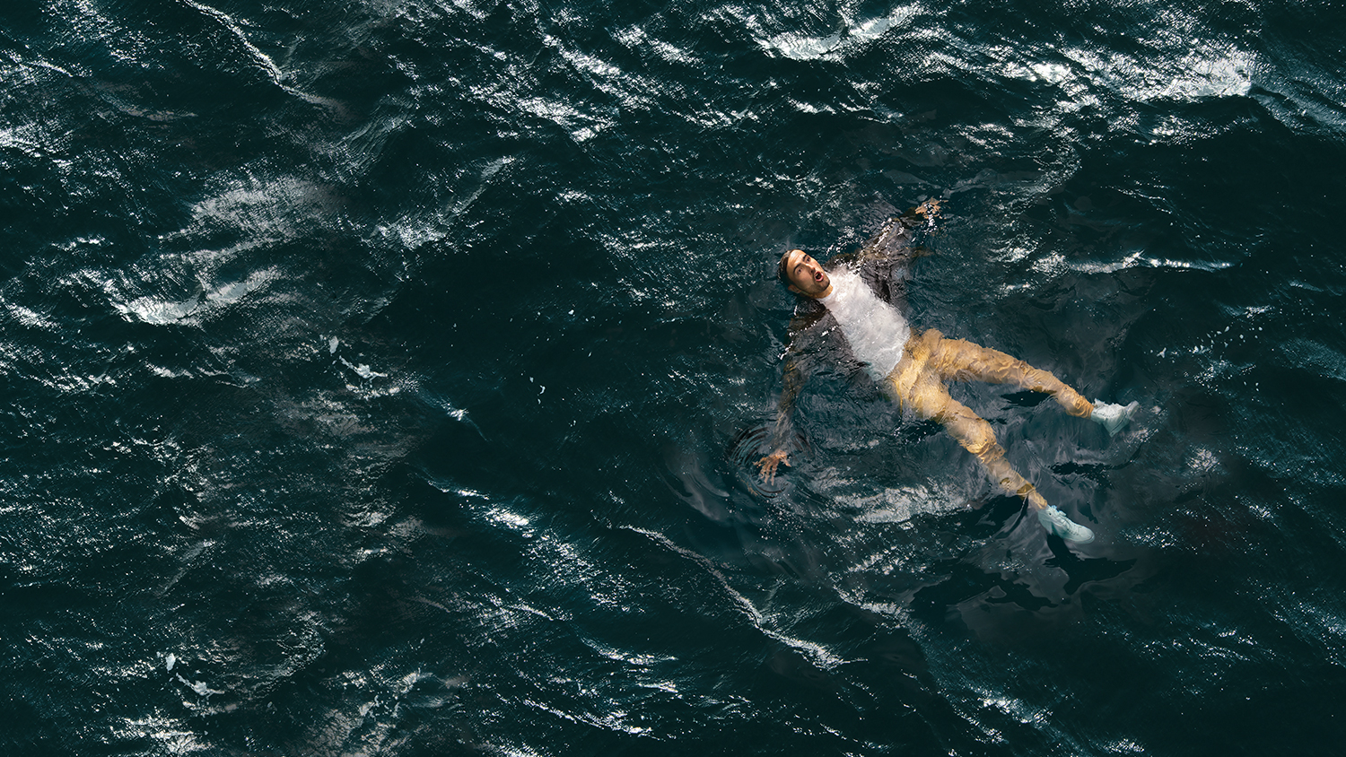 A man wearing a loose shirt, tshirt, trousers and shoes floating on his back, in the sea, with arms and legs