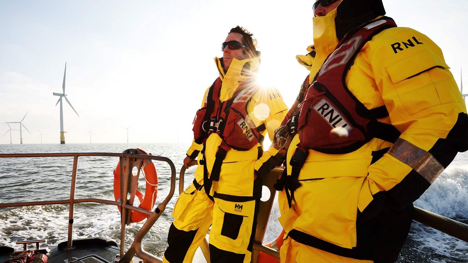 Two crew members onboard a Shannon class lifeboat wearing Helly Hansen all-weather crew kit. The sun is beating down on the lifeboat and there is a wind farm in the background.
