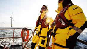 Two crew members onboard a Shannon class lifeboat wearing Helly Hansen all-weather crew kit. The sun is beating down on the lifeboat and there is a wind farm in the background.