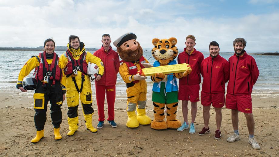 The RNLI’s Stormy Stan and Haven’s Rory the Tiger standing with two lifeboat volunteers, two members of RNLI staff and a Haven guest with her dog on the beach with the sea in the background 
