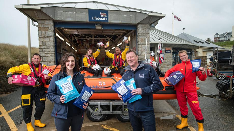 Bude’s D class lifeboat on its trailer outside the boathouse with two lifeboat volunteers sitting inside, one standing next to it on the left, an RNLI lifeguard standing to its right, and Lucy Ashton from the RNLI and Dave McNulty from Burts Snacks standing in front of it. All smiling and holding packs of RNLI-branded Burts Snacks hand-cooked crisps. 