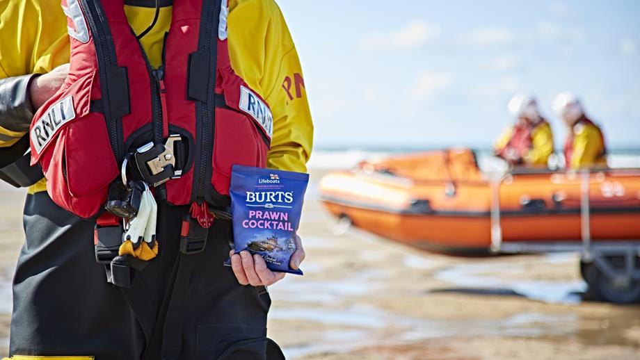RNLI Crewman standing on beach holding a packet of Burts prawn cocktail flavour crisps