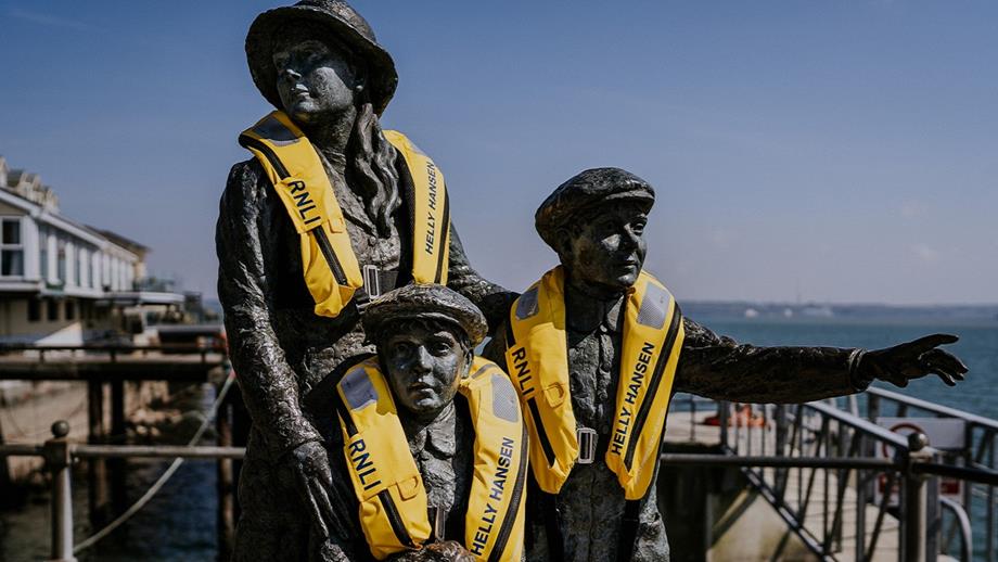 Three bronze figures, a statue of a mother and two children, wear bright yellow life-jackets with the blue sea in the background.