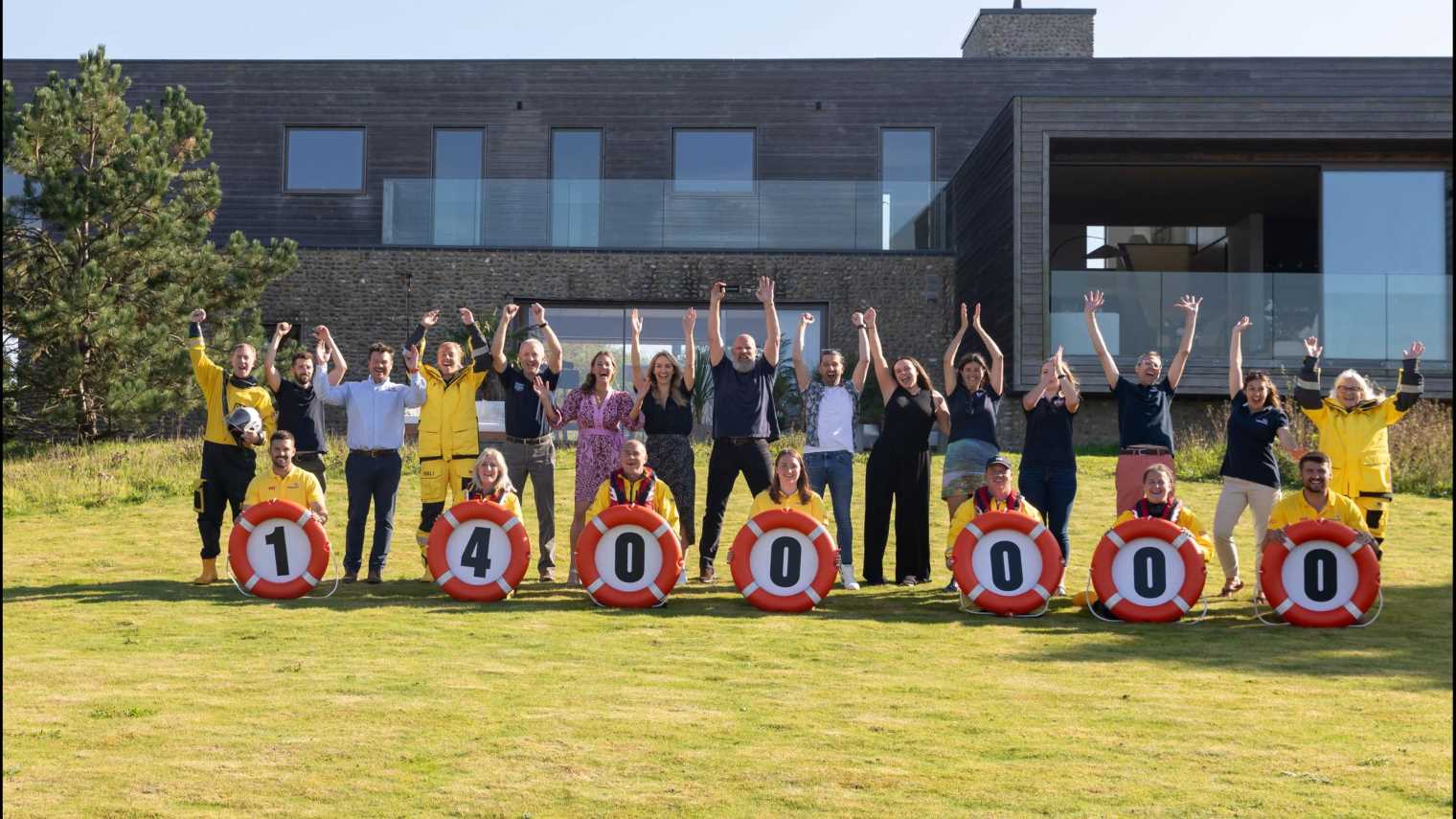 A line of people cheering with arms in the air in front of the luxury house in Norfolk, including Grand Prize winner Daren Bell. RNLI lifeboat volunteers and lifeguards are kneeling in front of them holding liferings with numbers inside showing the value of the house - £1,400,000. 