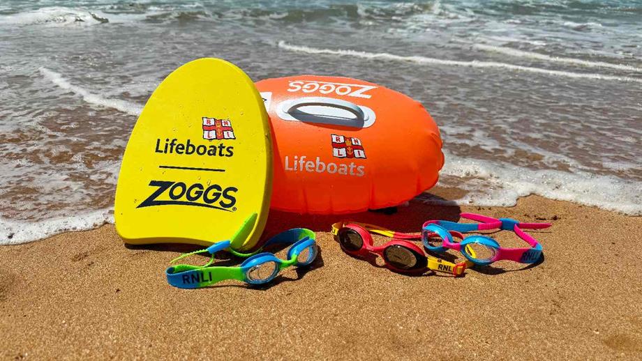 Zoggs kickboard, rubber ring and swimming goggles with RNLI branding displayed on a beach with waves coming in