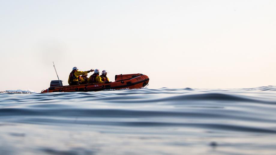 An inshore lifeboat is afloat on the horizon in relatively calm waters underneath a bright white cloudy sky.