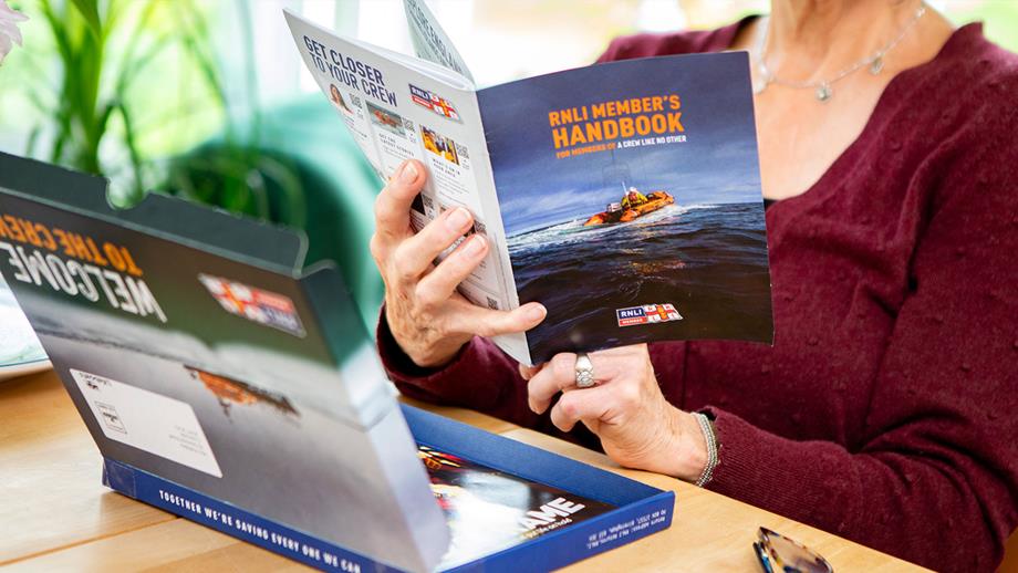 An RNLI member opens their welcome pack, with the box open on the table and the Member’s Handbook open in their hands