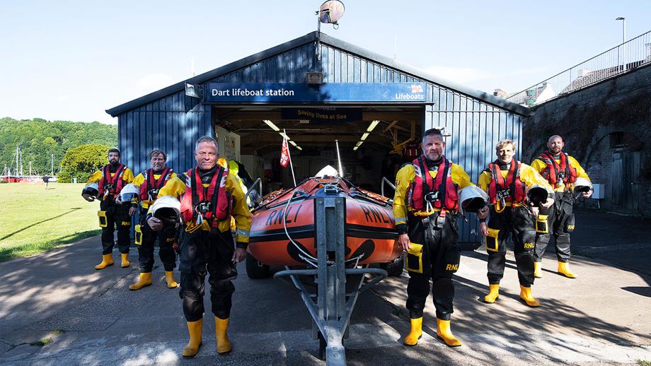 Six Dart lifeboat volunteers stood outside of the boathouse around the D class lifeboat, all wearing full crew kit