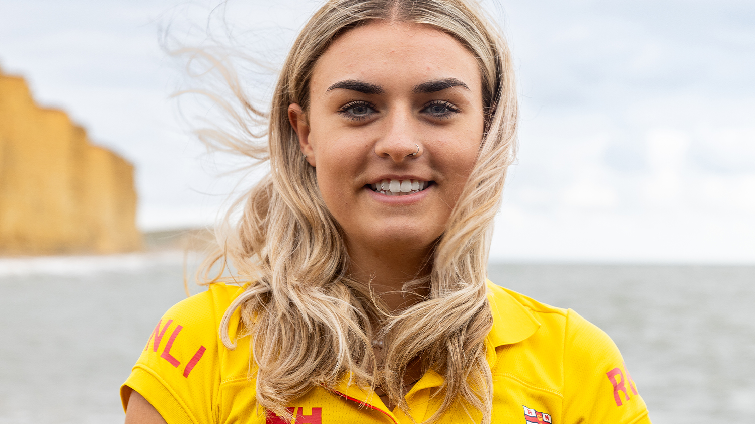 RNLI Lifeguard Abigail Greenslade smiles at the camera. She is wearing a yellow lifeguard polo and the sea is in the background.