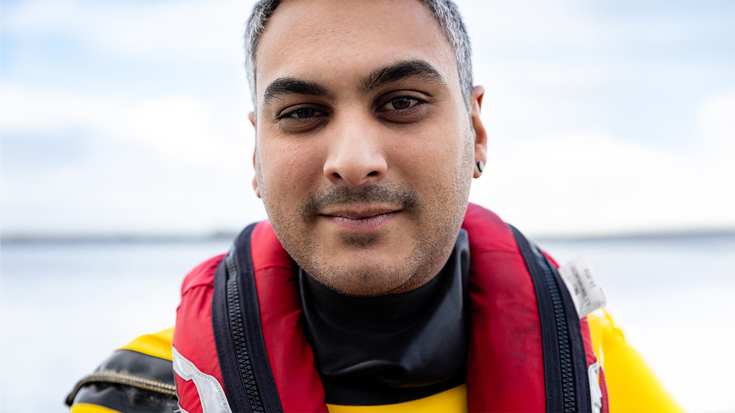 A portrait of Nathan Vasudaven, Volunteer Crew Member at Gravesend RNLI. He is wearing crew kit. Blue skies and the sea are in the background.