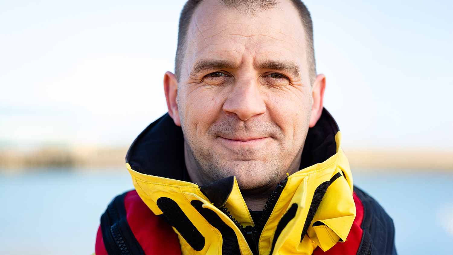 Simon Williams, Coxswain Mechanic at Shoreham Harbour Lifeboat Station, smiles at the camera. He is wearing crew kit. Blue skies and the sea are in the background.