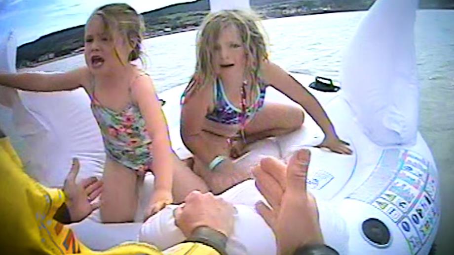 Hands reach out in front of the camera to rescue two young girls, who are crying. The girls are on an inflatable swan some distance from the shore