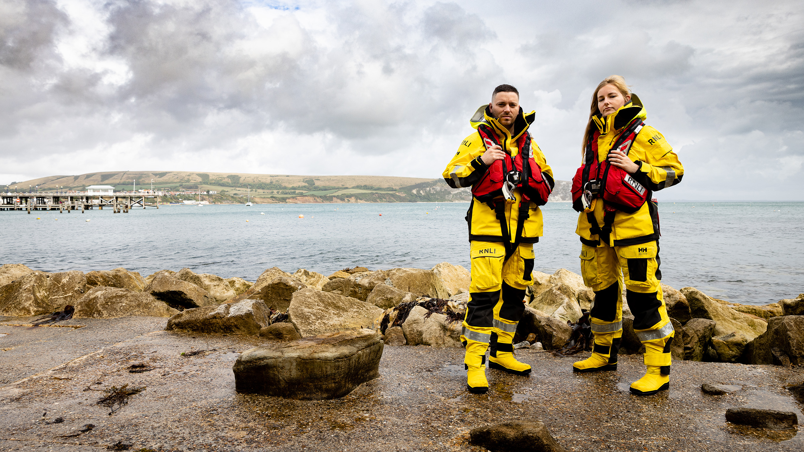 Two crew members standing on a rocky path with the sea behind them. They are looking into the camera.