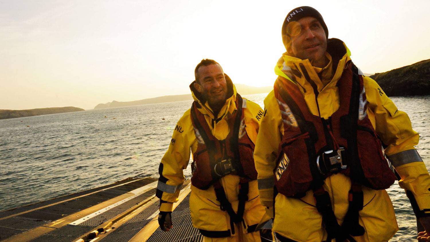 St Davids RNLI Crew Members Roger Smith and Dai John in their all-weather kit walking up the slipway as the sun sets in the distance