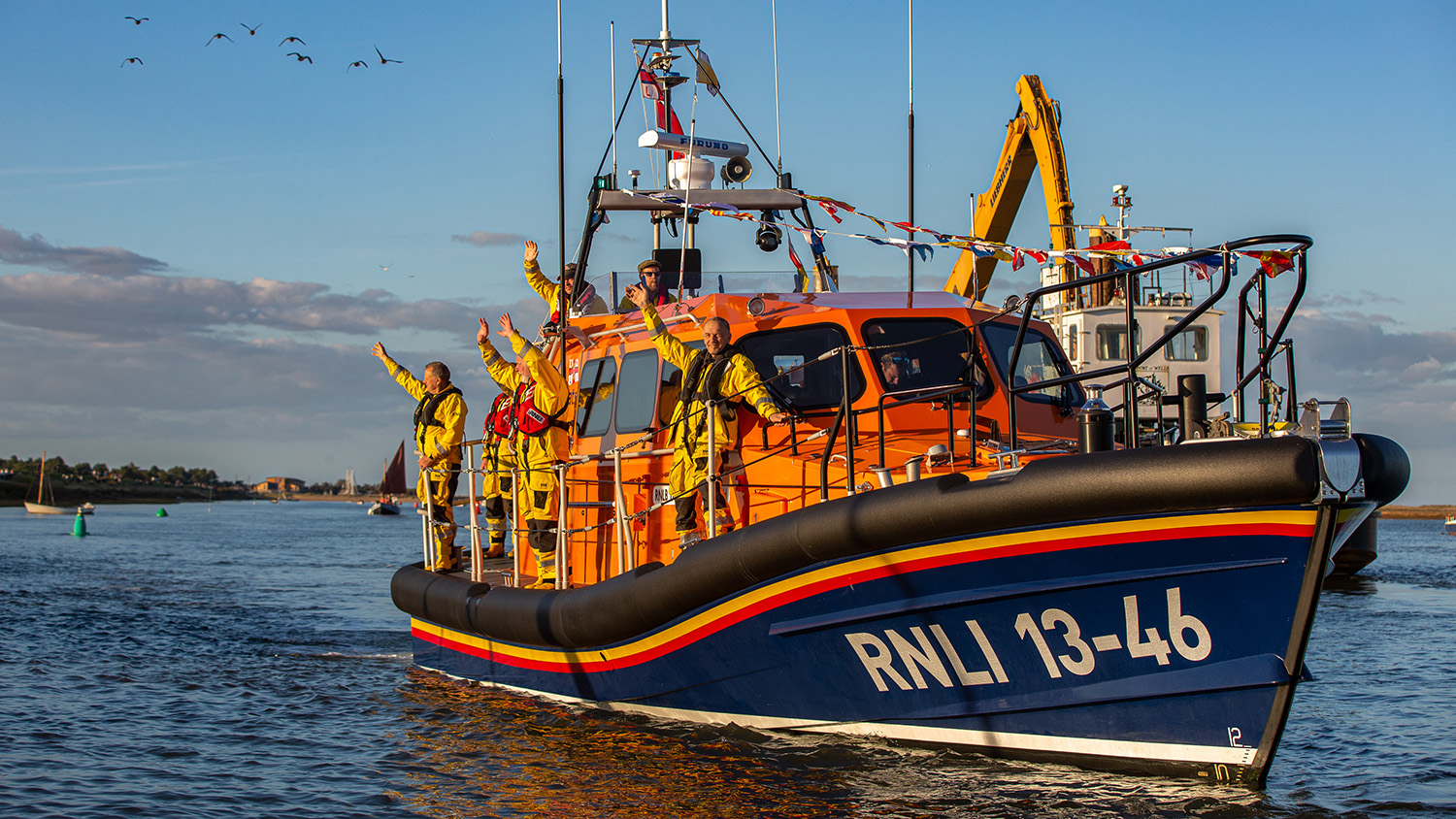 Crew stand waving on the deck of the Wells lifeboat which is decorated with bunting.