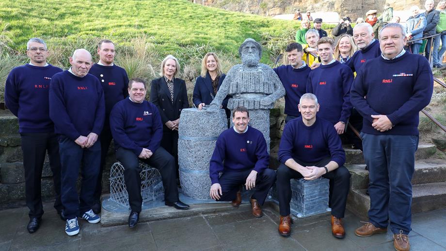 Whitby RNLI volunteers with the Henry Freeman sculpture, artist Emma Stothard and Lesley Jackson, a descendant of Henry, at Khyber Pass. A grassy bank and cliffs are in the background.