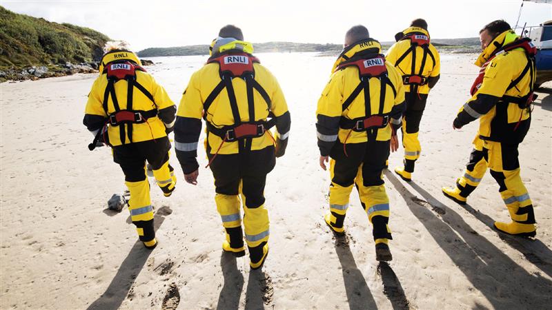 Five lifeboat volunteers in all-weather crew kit walking along the beach away from the camera at Clifden on a bright, sunny day