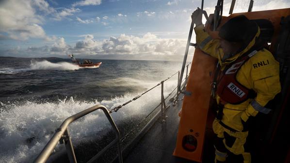 A volunteer at the stern of Clifden RNLI’s Shannon class all-weather lifeboat while at sea. The station’s B class inshore lifeboat crew are in the background. Sunny day. White spray around the lifeboats.