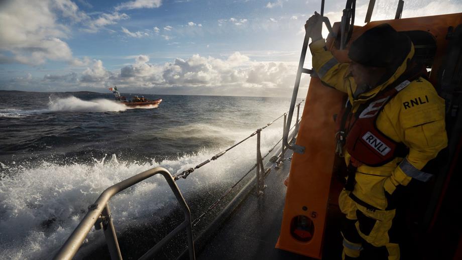A volunteer at the stern of Clifden RNLI’s Shannon class all-weather lifeboat while at sea. The station’s B class inshore lifeboat crew are in the background. Sunny day. White spray around the lifeboats.