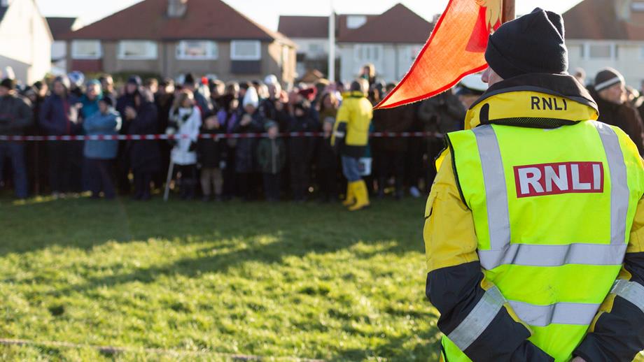 A man in a high-vis RNLI jacket has his back to the camera, as though an event is about to begin. Flags wave. A person in lifeboat crew kit is asking the watching crowd for donations to a collection bucket.