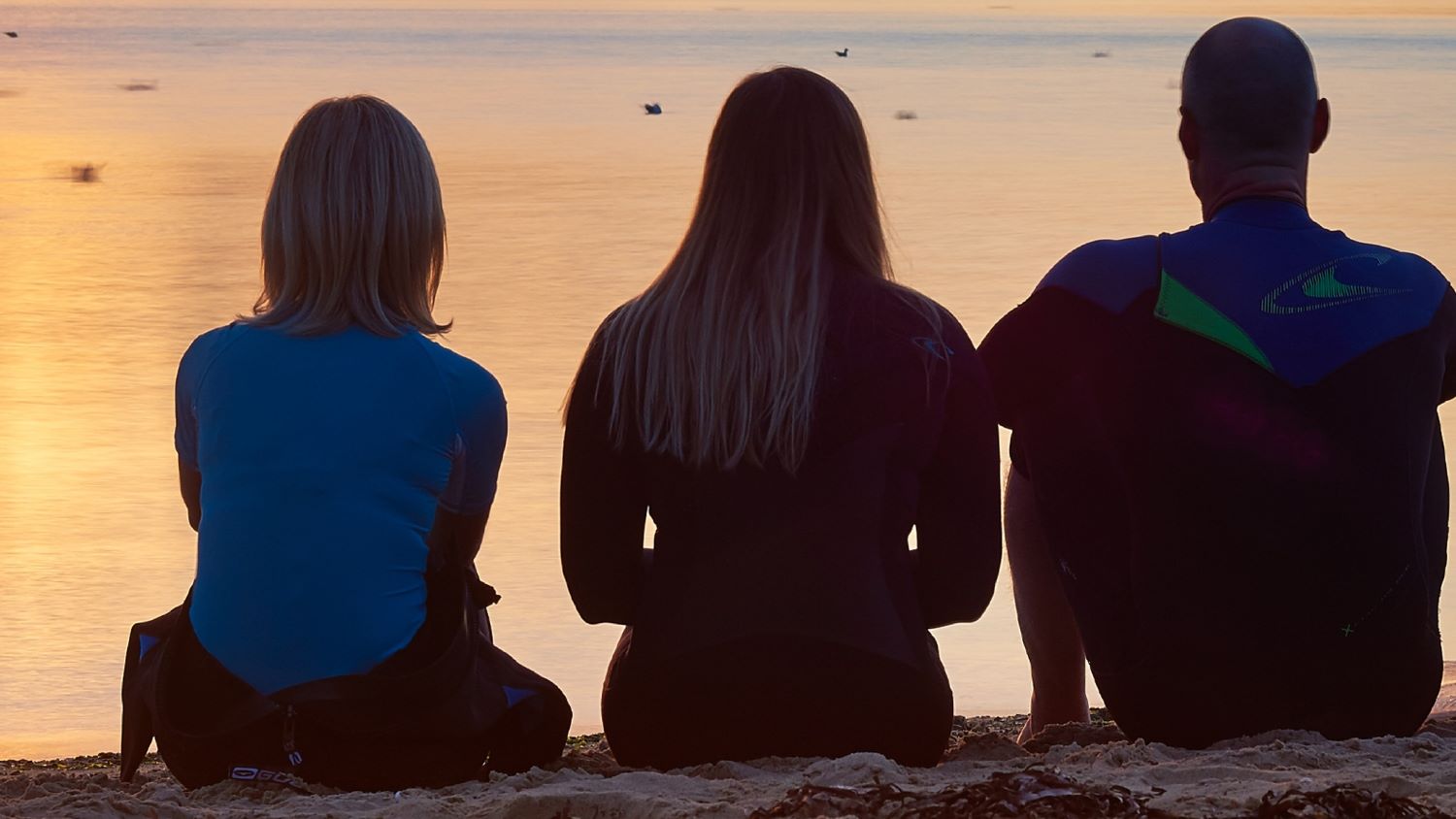 Three people sit on a beach with their backs to the camera, gazing over the water towards a beautiful sunset