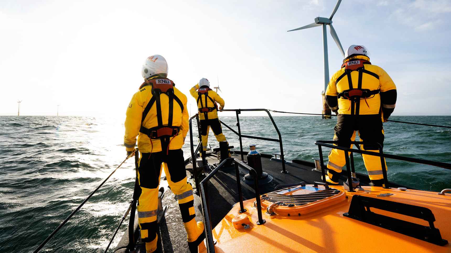 Three lifeboat crew members are stood on the bow of a Shannon class lifeboat, looking out to sea on a sunny day with wind turbines ahead of them.