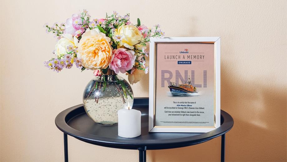 Close up photo of a picture of a Launch a Memory celebration certificate inside a photo frame on a table next to a lit candle and a vase with flowers in it