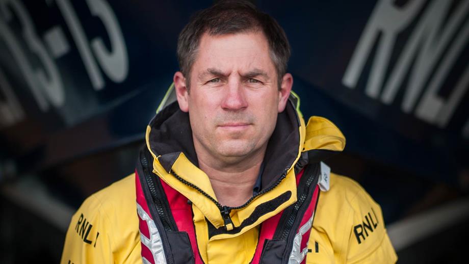Portrait of Swanage Lifeboat Coxswain Dave Turnbull. Dave stands in front of the Shannon class lifeboat wearing all-weather lifeboat kit and a lifejacket