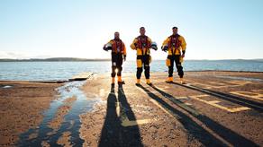 3 male RNLI crew members stand next to a body of water