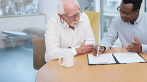 A senior man with white hair and glasses sits at a table alongside a younger man with glasses, wearing a light blue shirt. The younger man is explaining a document on a clipboard, gesturing with a white pen, while the older man listens attentively. They are sat next to each other in a modern office, with white coffee mugs on the table. 