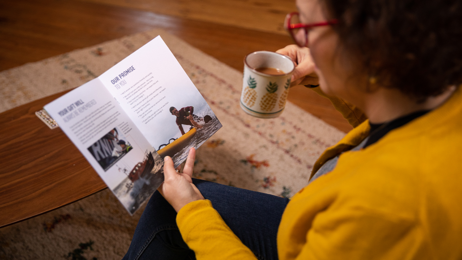 A woman reading the RNLI’s free guide to gifts in Wills with a cup of tea. There is a coffee table and rug on top of a wooden floor. She has short brown hair and is wearing a yellow cardigan over a dark top, dark jeans and dark-rimmed glasses.  