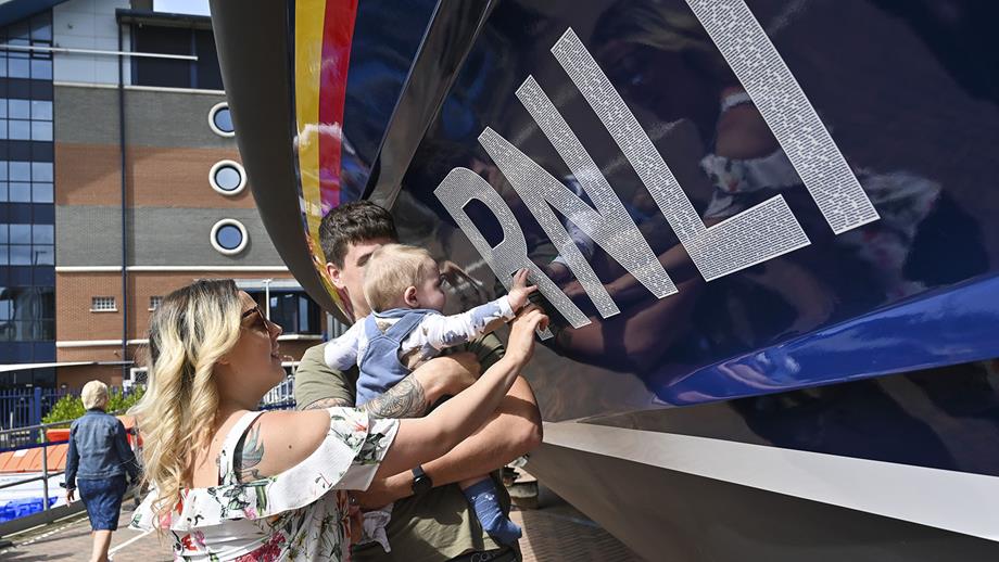 A woman, a man and a baby are standing next to the hull of a Shannon class lifeboat and are looking and pointing at the decal.