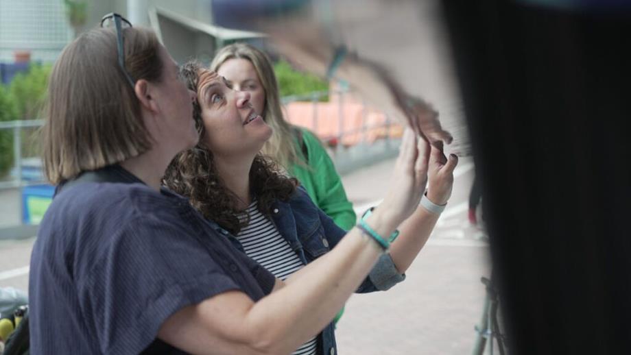 Two women are standing next to the lifeboat. They are looking up at the lifeboat and pointing to find the names of their loved ones. The woman closest to the camera has short straight hair and is wearing a navy blue short-sleeve shirt. The woman standing next to her has brown curly hair, and is wearing a striped top with a denim jacket. There is another woman in the background wearing a green top.