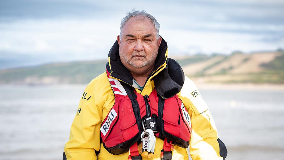 Portrait photo of Dan Potter, Volunteer Coxswain at New Quay Lifeboat Station, outside in all-weather lifeboat kit with the sea and coastline in the background.