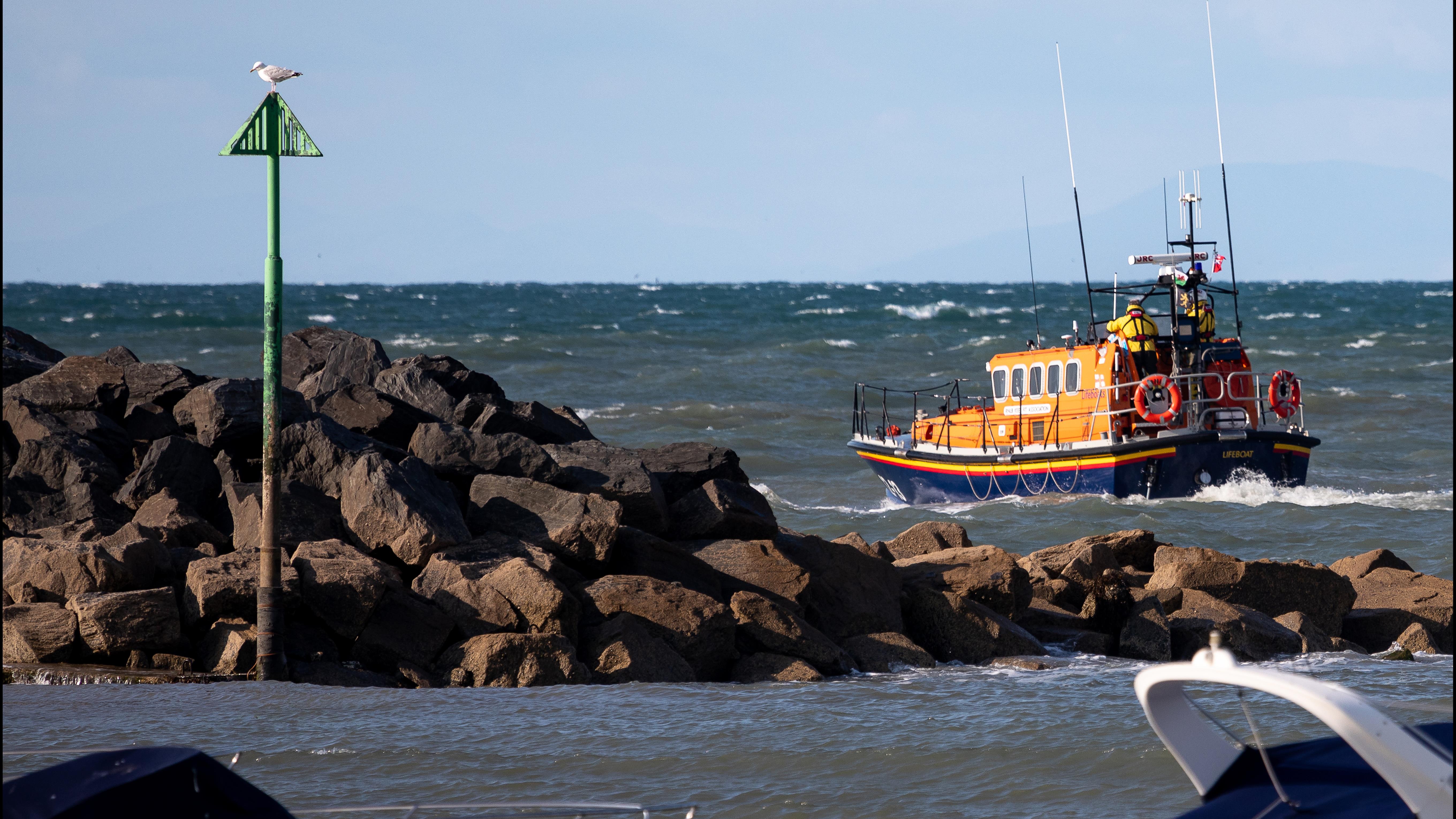 Photo taken from the shore of New Quay lifeboat crew on a training exercise onboard relief Mersey class all-weather lifeboat Keep Fit Association 12-13. The lifeboat is just passing the rock armour of New Quay Harbour and the back of two lifeboat members can be seen at the stern. Clear blue skies and choppy water.