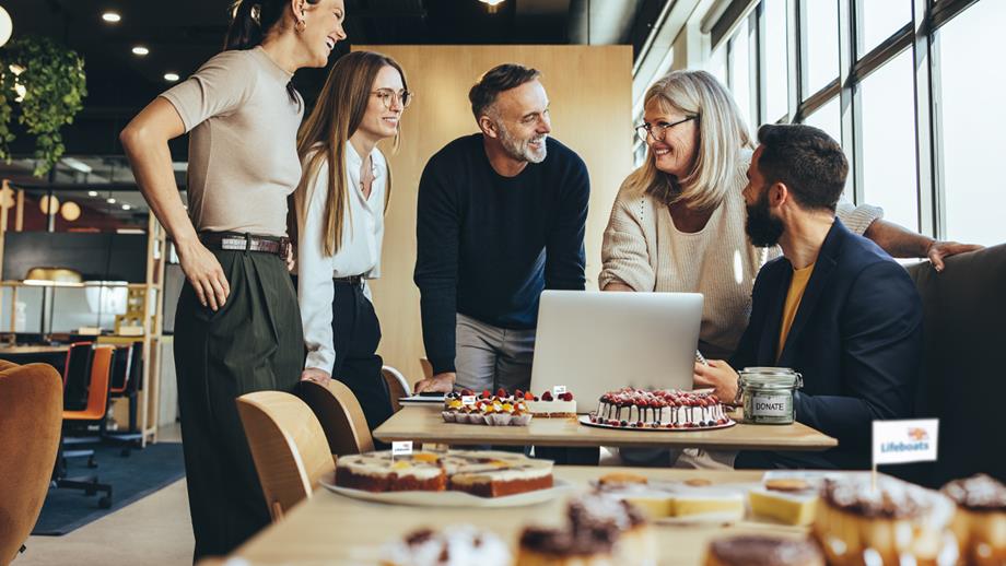 Five smiling people are in an office, standing behind tables full of cakes that are being sold to raise money for charity