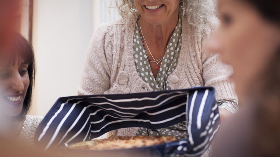 A woman is holding a dish full of food with oven gloves and smiling.