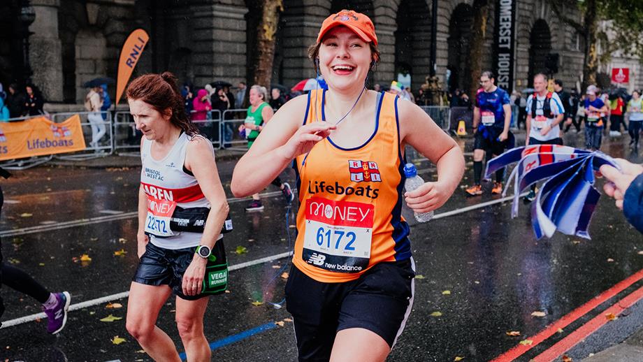 A smiling RNLI runner on a wet city street, with other runners around them. They smile to supporters off camera