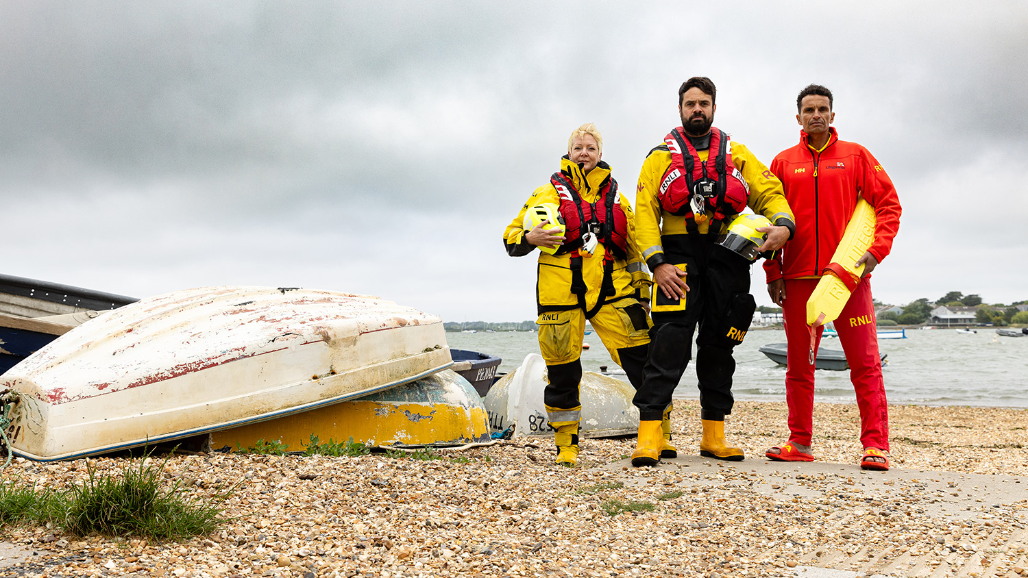Two lifeboat crew members and an RNLI lifeguard are stood next to small upturned boats on a beach on a cloudy day. 