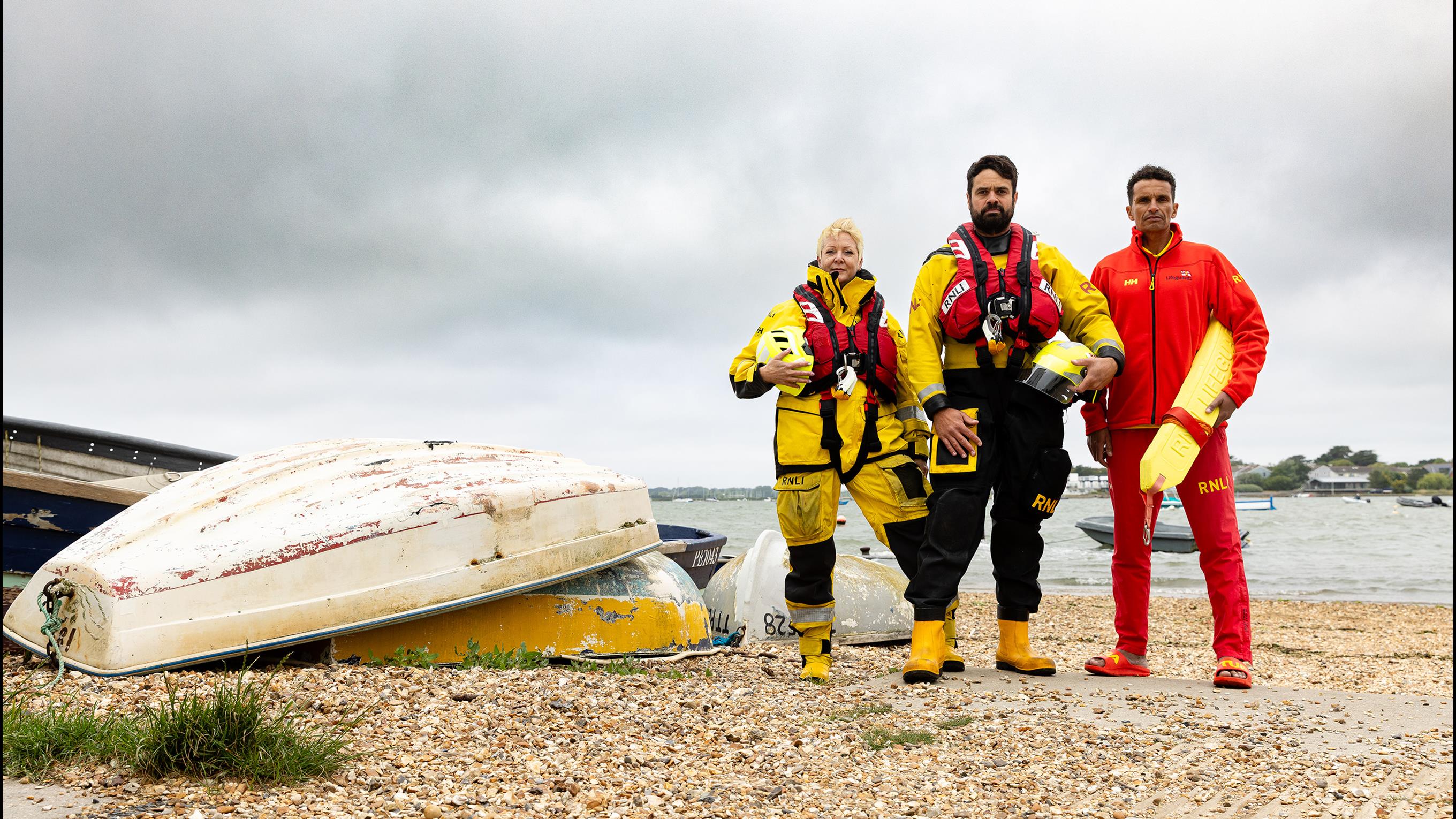 Two lifeboat crew members and an RNLI lifeguard are stood next to small upturned boats on a beach on a cloudy day. 
